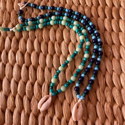 Três colares boémios de contas coloridas com pendentes de conchas em fundo texturizado castanho.