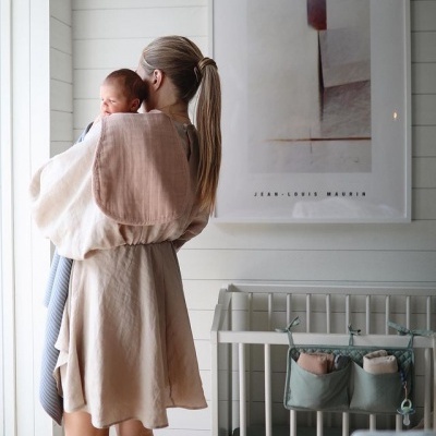 Mulher segurando bebé enrolado em mantas de algodão junto a berço branco num quarto minimalista