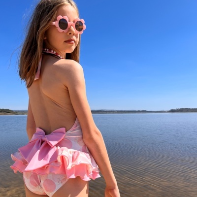 Menina com fato de banho rosa e óculos de sol flor junto a lago sob céu azul