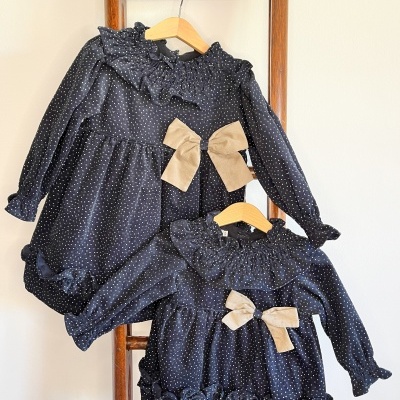 Dois vestidos infantis pretos com pontos brancos, folhos e laços bege pendurados em cabides numa escada de madeira