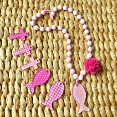 Colar rosa com peixe de madeira e conta bolinhas, peixes e cruzes de madeira rosa sobre tecido trançado.