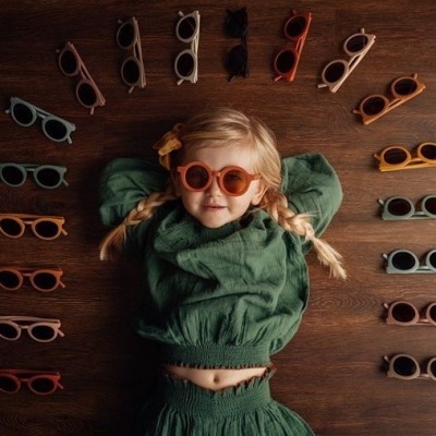 Criança com óculos de sol laranja e roupa verde, rodeada por vários óculos de sol coloridos em círculo sobre madeira escura