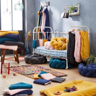 Quarto com cama branca em metal e almofadas coloridas em tons azul, amarelo e rosa, com roupa pendurada e têxteis dobrados em tons vibrantes.