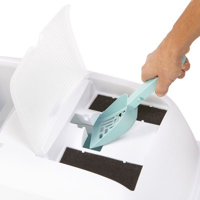 Caixa de areia branca com tampa aberta e uma mão a usar pá verde-água no interior