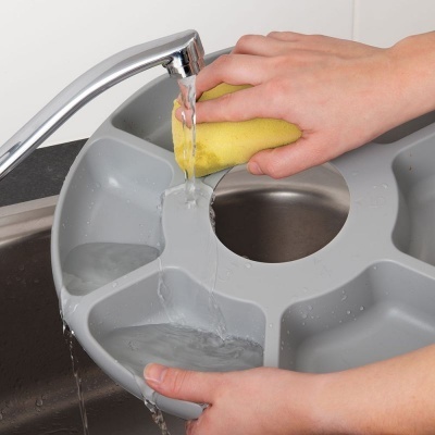 Tabuleiro cinzento de plástico com divisórias e abertura central a ser lavado numa torneira.