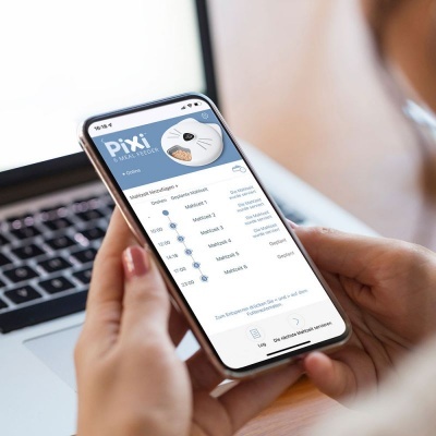 Pessoa a segurar um smartphone a mostrar uma aplicação de medicação chamada PIXI com gráfico de doses, em frente a um computador portátil.