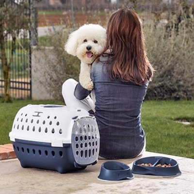 Caixa transportadora de cão branca e azul com dois recipientes azuis e cão branco ao colo de mulher sentada ao ar livre