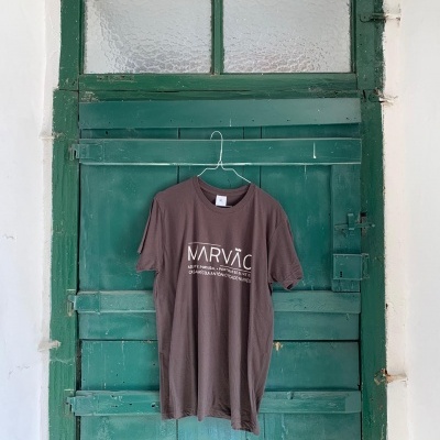 Camiseta castanha com texto branco pendurada numa porta verde de madeira com vidro fosco