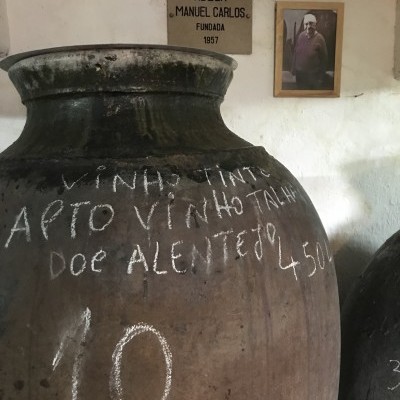Ânfora de barro com escrita a giz e parede com placa e fotografia ao fundo