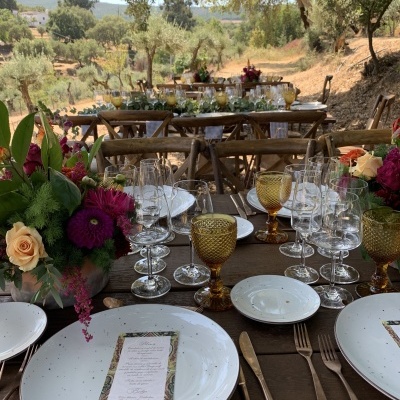 Mesa rústica ao ar livre com louça branca, copos transparentes e amarelos, talheres dourados e flores coloridas