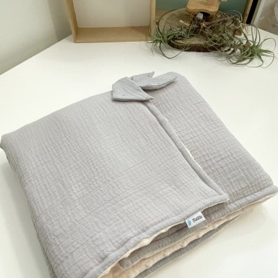 Manta infantil cinzenta de algodão dobrada sobre mesa branca com decoração de madeira e plantas.