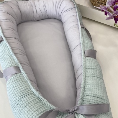 Ninho de bebé cinzento e verde-água com padrão waffle e fita cinzenta, etiqueta Catita, caixa com palavras CATITA KIDS e flores.