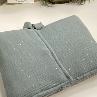 Almofada azul claro com pontos dourados e etiqueta Catita sobre mesa branca com planta e quadro ao fundo