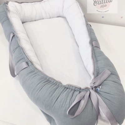 Ninho para bebé cinzento e branco com fitas, sobre uma superfície branca, parede com pontos coloridos e logotipo Catita Kids.