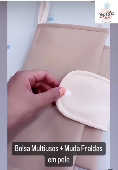 Bolsa multiusos bege com muda fraldas creme, textura de pele, fecho em velcro, sobre superfície branca com pontos azuis.