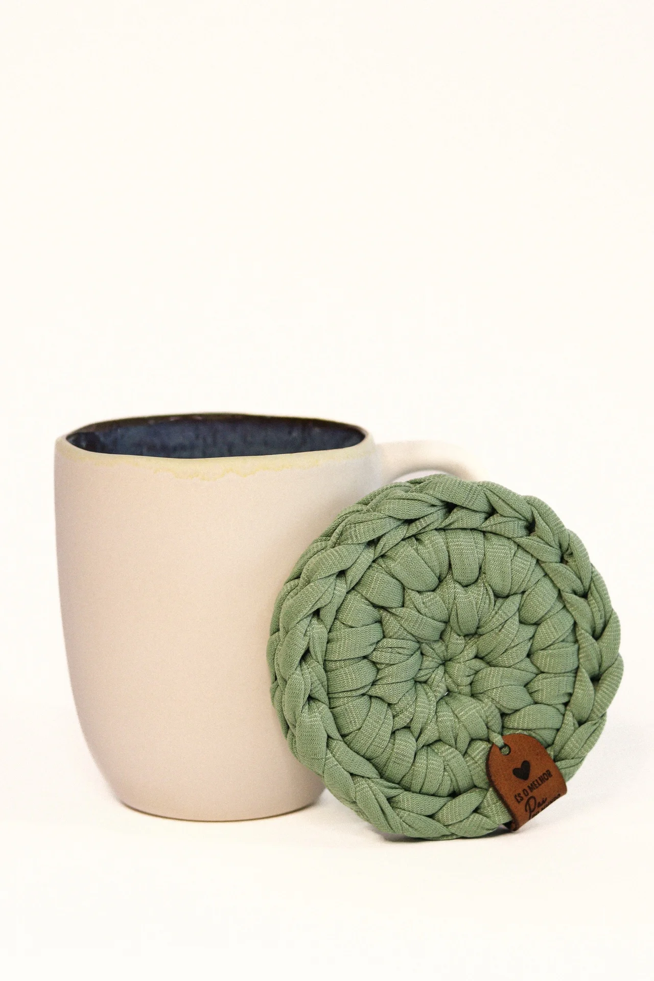 Caneca bege com interior azul e apoio verde trançado.