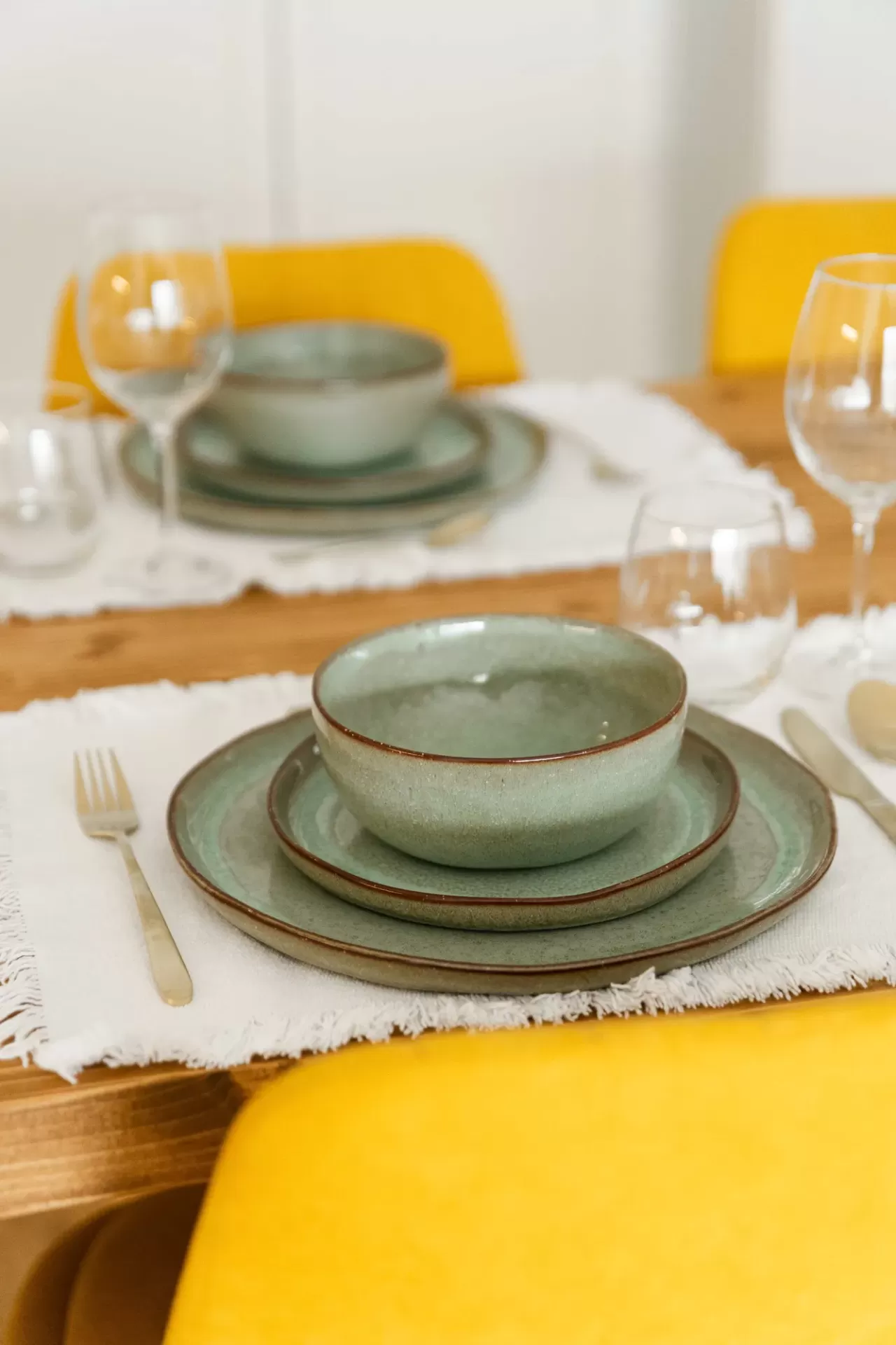 Conjunto de loiça verde-azulado com pratos e tigela sobre mesa de madeira com toalha branca e cadeiras amarelas