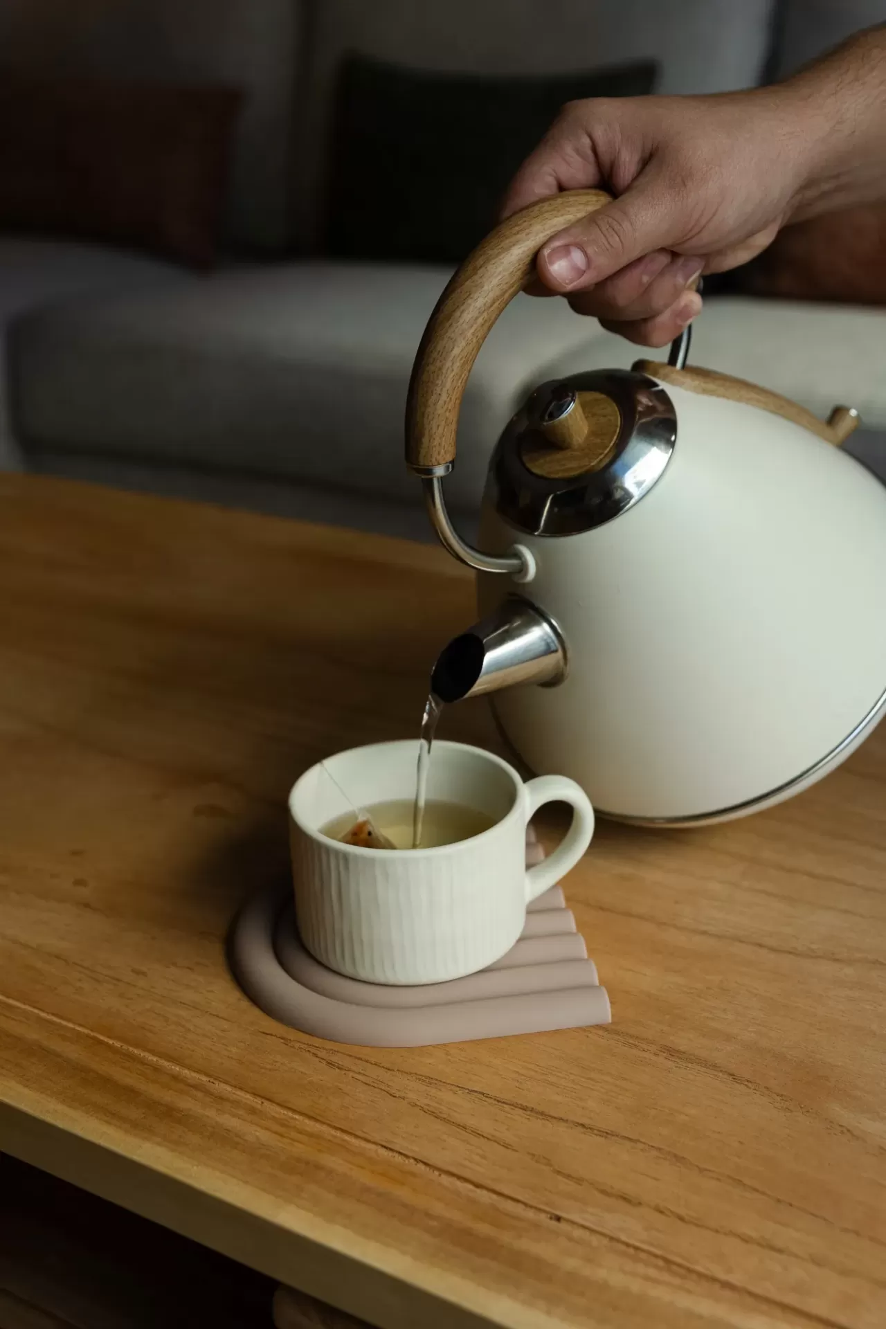 Bule branco com pega de madeira a verter água para chávena em mesa de madeira.