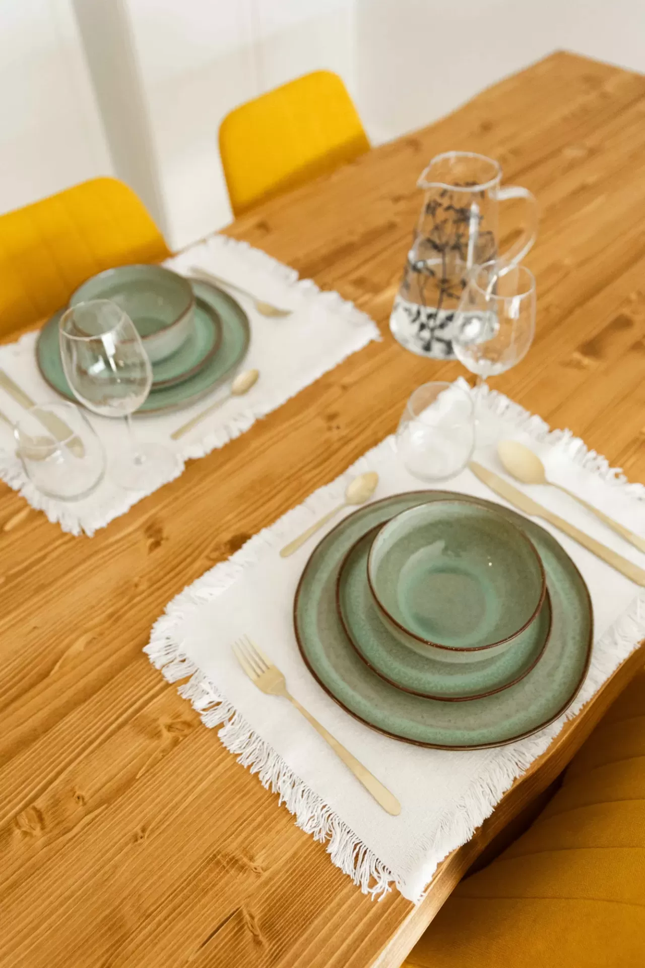 Mesa de madeira preparada para refeição com pratos verdes, talheres dourados, taça de vinho, jarra de vidro decorada e cadeiras amarelas.