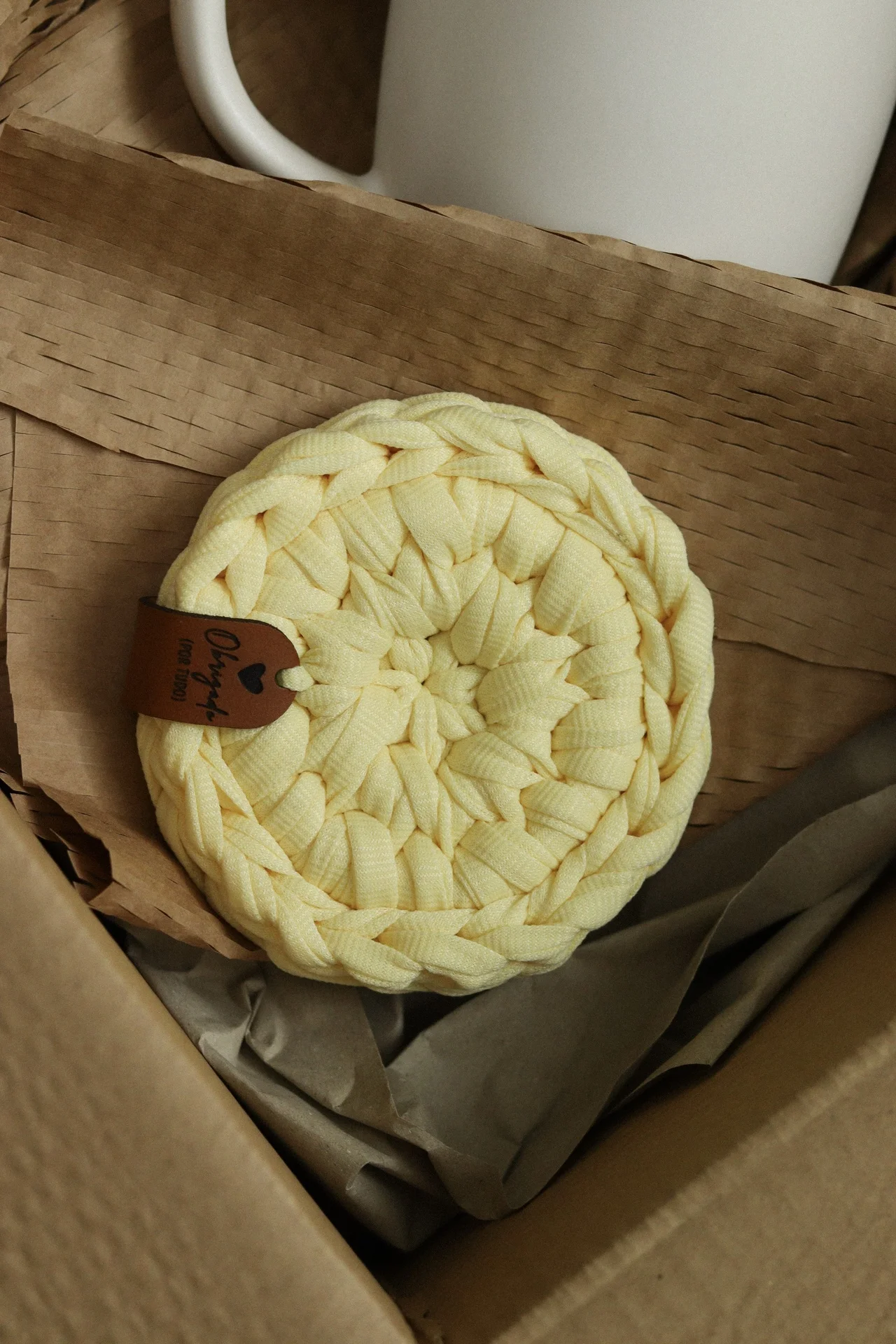 Descanso de panela amarelo trançado com etiqueta de couro dentro de caixa de papelão.
