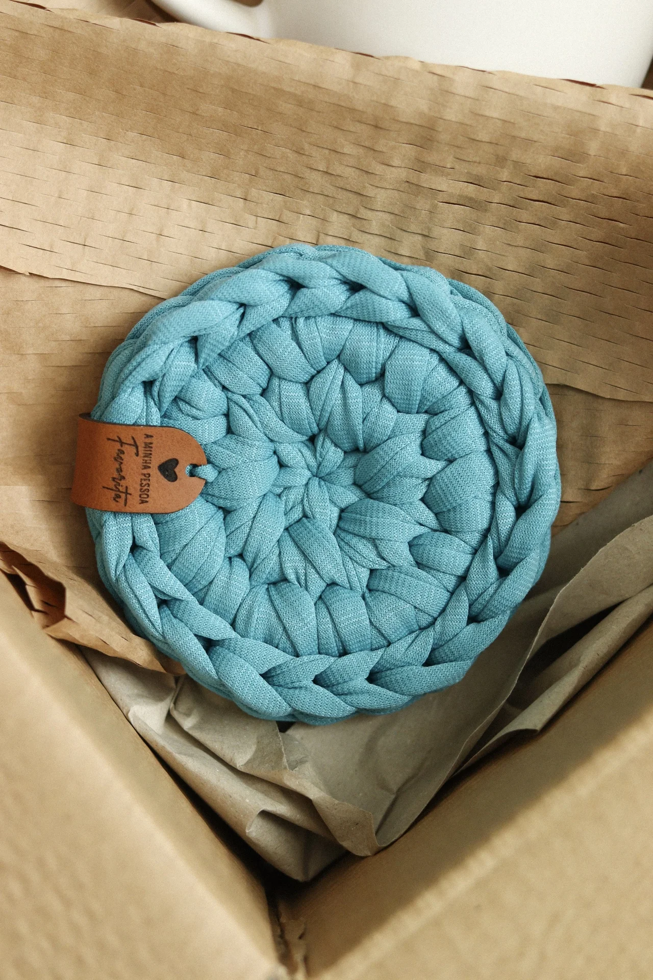 Descanso de panela tricotado azul claro com etiqueta castanha dentro de caixa de papelão
