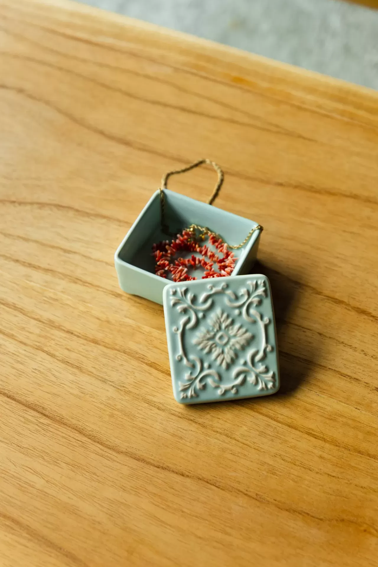 Caixa de cerâmica azul clara com tampa ornamental e colar vermelho dentro, sobre mesa de madeira.
