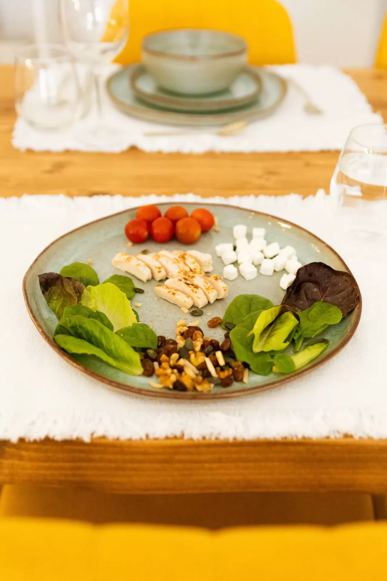 Prato com salada, tomates, frango, queijo e nozes sobre mesa de madeira com cadeiras amarelas