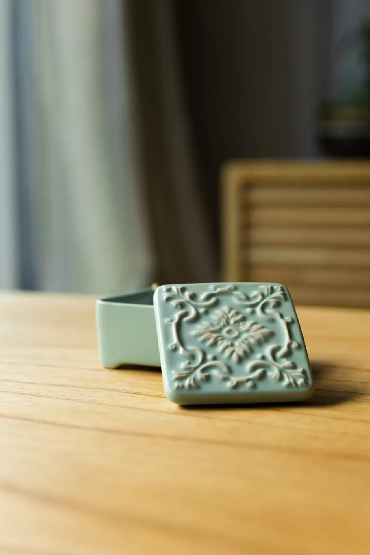 Caixa quadrada de cerâmica verde-água com tampa decorada com padrão em relevo numa mesa de madeira.
