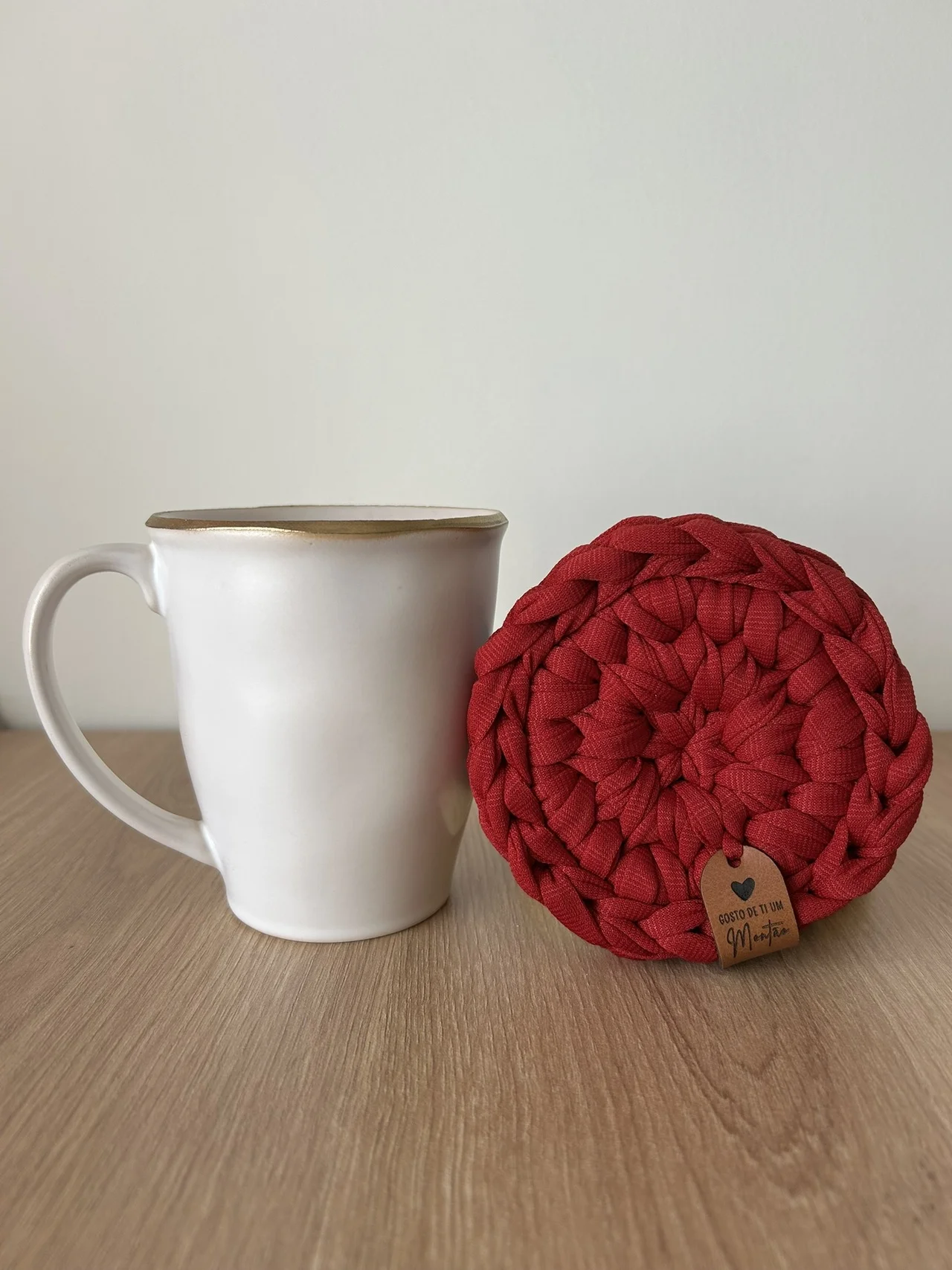 Caneca branca com borda dourada ao lado de suporte vermelho trançado com etiqueta de couro sobre mesa de madeira