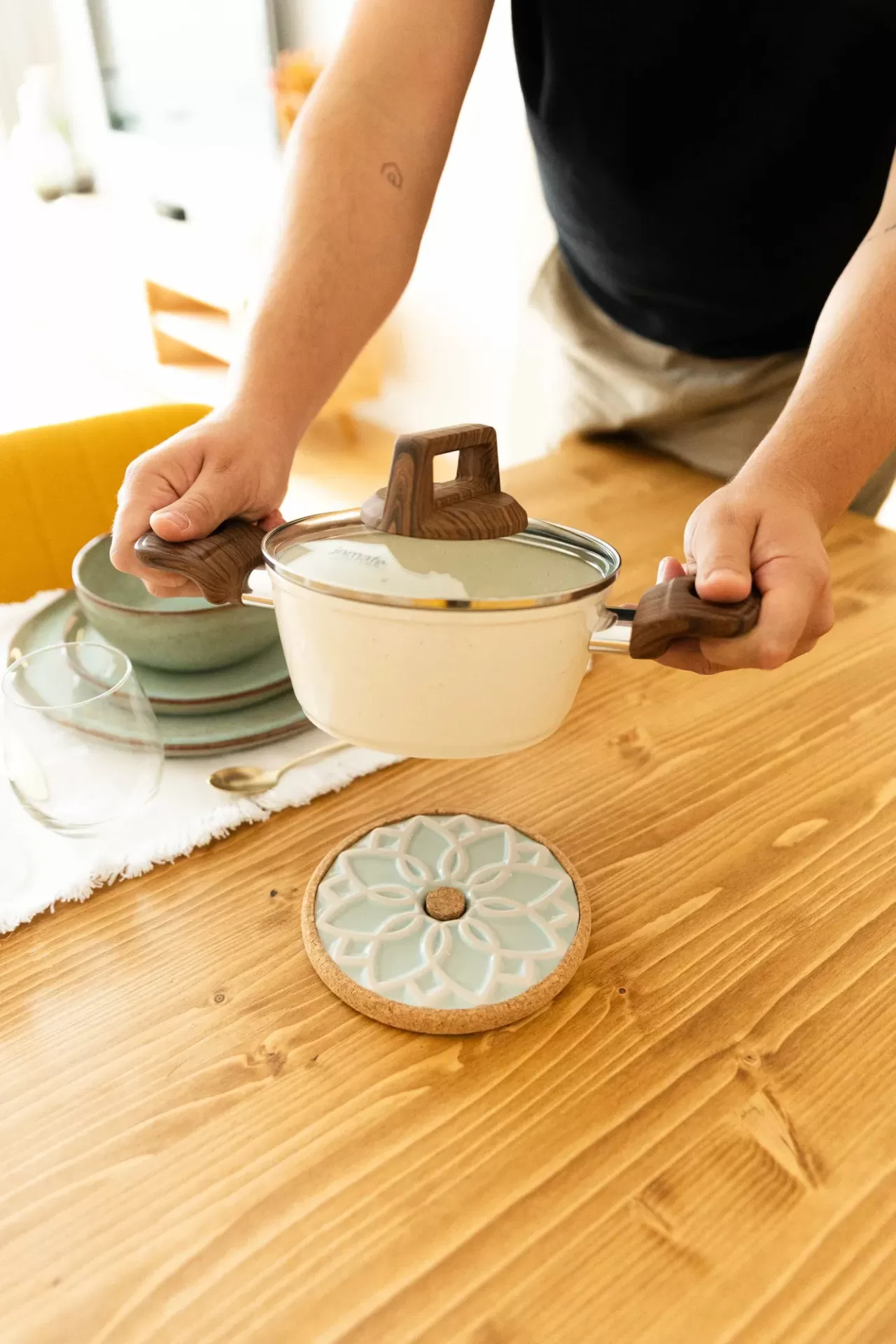 Panela branca com pega em madeira e tampa de vidro sobre suporte decorativo verde e castanho numa mesa de madeira.