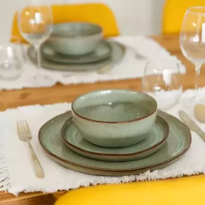 Conjunto de loiça verde-azulado com pratos e tigela sobre mesa de madeira com toalha branca e cadeiras amarelas