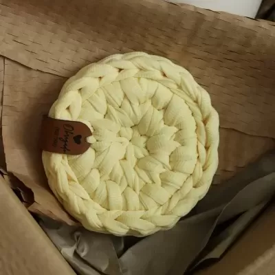 Descanso de panela amarelo trançado com etiqueta de couro dentro de caixa de papelão.