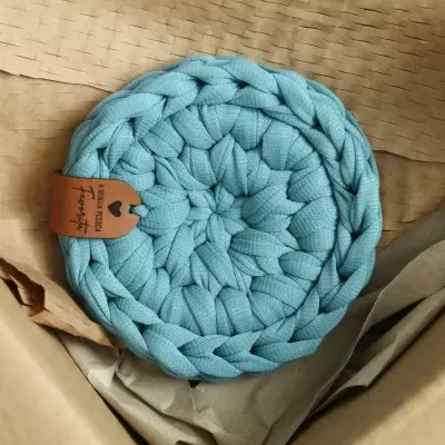 Descanso de panela tricotado azul claro com etiqueta castanha dentro de caixa de papelão