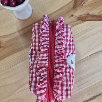 Estojo xadrez vermelho e branco com fecho vermelho e babados, sobre mesa de madeira clara com taça branca de cerejas.
