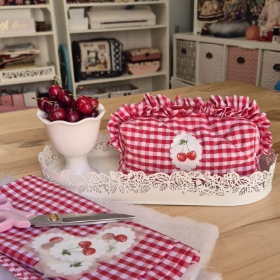 Bolsa almofadada xadrez vermelho e branco com cerejas, tesoura, tecido e cerejas frescas sobre mesa de madeira em sala com estantes e bonecas