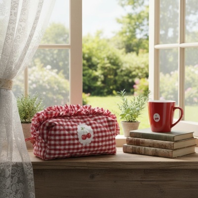 Cesta de tecido vermelho e branco com padrão xadrez e detalhe de cerejas, caneca vermelha com adesivo de cereja, livros empilhados, vasos com plantas na janela