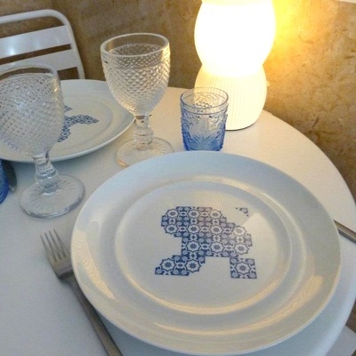 Mesa posta com pratos brancos com padrão azul, copos de vidro transparentes e azuis, talheres e lâmpada acesa