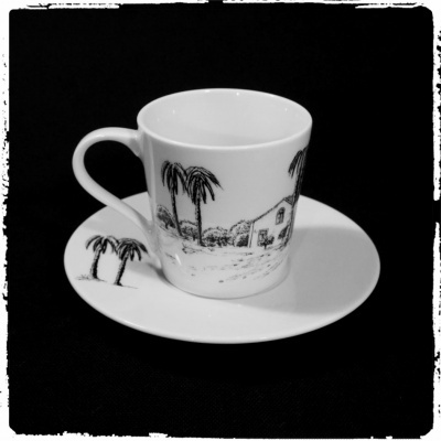 Chávena e pires brancos com desenhos a preto de palmeiras e casa