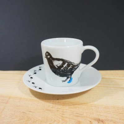 Copo branco com desenho de pássaro e pires com padrão preto sobre madeira clara
