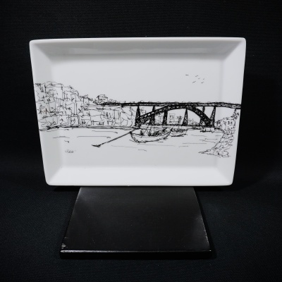 Prato rectangular branco com desenho preto de paisagem com ponte e barcos.