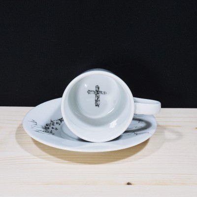 Chávena branca com cruz preta no interior e prato com padrão preto numa superfície de madeira clara