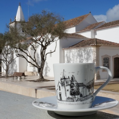 Chávena e pires de porcelana branca com desenho a preto de igreja, em frente a edifício branco com telhado e árvores