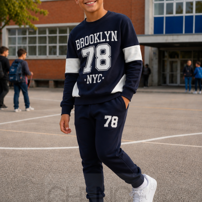 Rapaz com conjunto azul escuro com texto BROOKLYN 78 NYC e ténis brancos num pátio escolar.