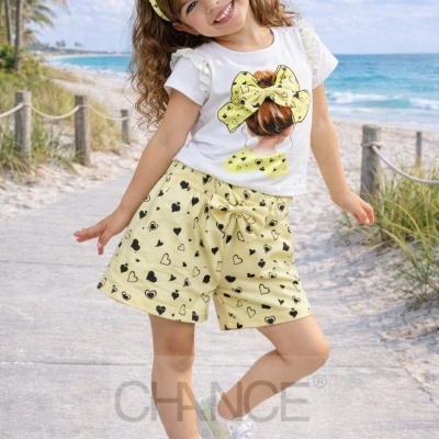 Menina criança vestindo conjunto amarelo com corações pretos e t-shirt branca com desenho, posando numa passadeira à beira da praia.