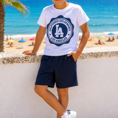 Menino de camiseta branca com logotipo azul, calções azuis e sapatilhas brancas junto à praia e mar.