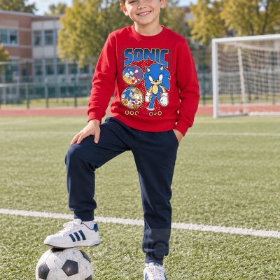 Criança com sweat vermelho estampado Sonic, calças azuis e ténis brancos num campo de futebol.