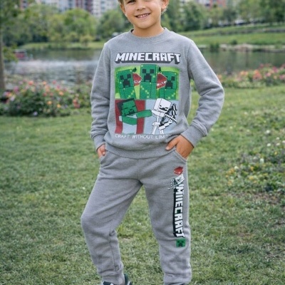 Criança a usar conjunto cinzento Minecraft com personagens verdes e brancos, em parque com relva e prédios ao fundo.