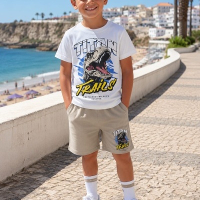 Menino sorridente com t-shirt branca com dinossauro e calções bege com estampa semelhante, em passeio junto ao mar.