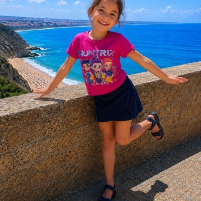 Menina sorridente com t-shirt rosa, saia azul e sandálias pretas perto do mar