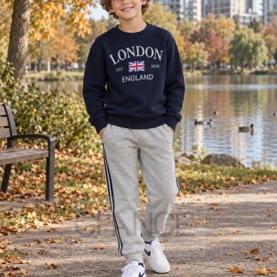 Criança de roupa casual com sweatshirt azul escuro, calças cinza e ténis brancos em cenário de parque outonal junto a lago.